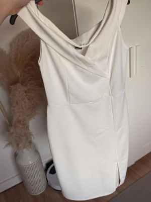 studentklänning från Gina Tricot - Säljer en stilren vit kortklänning från Gina Tricot med omlottdetalj vid halsen och en liten slits framtill. Klänningen är ärmlös och har en figurnära passform, perfekt för dig som gillar minimalistisk och modern stil.