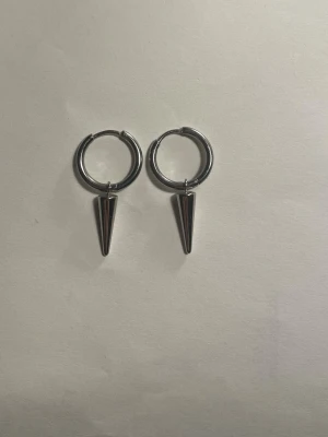 Edblad örhängen - Säljer ett par trendiga silvriga örhängen från Edblad med rund hoop och hängande konformad berlock. Örhängena har en modern och edgy look, perfekta för att lyfta din stil. Tillverkade i metall med blank finish. Andvända fåtal gånger, kommer såklart tvättade