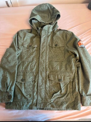 Grön jacka med tryck och luva - Snygg grön vårjacka med huva och coolt vitt tryck på ryggen där det står 'Chrome Rollers'. Jackan har fyra fickor framtill med knappar och ett patch på ena ärmen. Materialet ser ut att vara lätt och vindtätt, perfekt för blåsiga dagar.