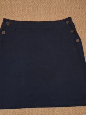 Mörkblå kortkjol med knappar - Mörkblå kortkjol i bomull med fyra dekorativa knappar på varje sida framtill. Kjolen har en rak passform och clean look.