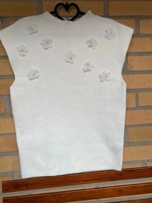 Vit stickad väst med blommor - Vit stickad väst med dekorativa blommor framtill, där varje blomma har en pärldetalj i mitten. Västen har en rak och något loose passform och är ärmlös. Perfekt att bära över en skjorta eller t-shirt för en söt och trendig look.