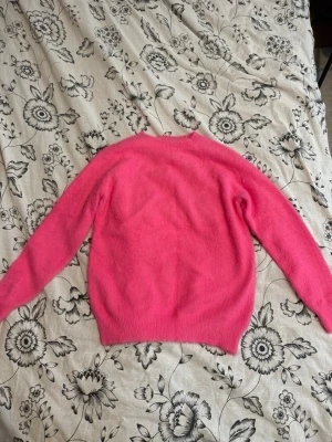 Mjuk rosa stickad tröja - En fluffig och mysig stickad tröja i stark rosa färg. Tröjan har rund halsringning, långa ärmar och ribbstickade muddar vid ärmslut och nederkant. Perfekt för dig som vill ha en färgklick i garderoben.