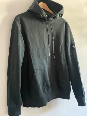 Mörkgrön hoodie från Calvin Klein Jeans - Snygg mörkgrön hoodie från Calvin Klein Jeans med stor CK-logga på ärmen. Storlek M.Hoodien har huva med dragsko, magficka och ribbade muddar vid ärmslut och nederkant. Tillverkad i mjuk bomull för skön känsla och avslappnad stil.