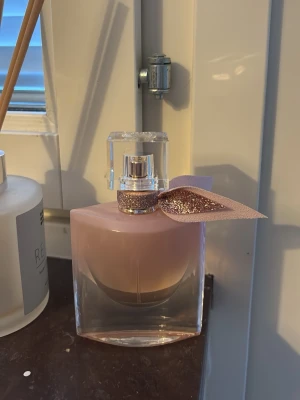 La Vie Est Belle parfym från Lancôme - Flaskan är helt full, har endast sprayat för att känna doften. Elegant parfymflaska från Lancôme, La Vie Est Belle, med mjukt rosa innehåll och transparent glas. Flaskan har en glittrig rosa rosett vid halsen och ett stilrent, genomskinligt lock. Perfekt som en lyxig detalj på sminkbordet. 
