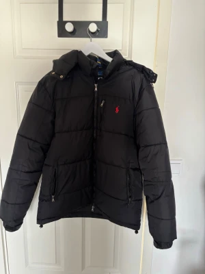 Svart pufferjacka från Ralph Lauren - Säljer en svart pufferjacka från Ralph Lauren med huva och dragkedja framtill. Jackan har två sidofickor med dragkedja