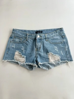 Ljusa blå jeansshorts med slitningar och Lågmidja  - Säljer ett par ljusa blå jeansshorts med låg midja och råa kanter och stora slitningar framtill. Klassisk femficksmodell med knapp och dragkedja. Perfekta för sommaren och ger en avslappnad vibe.