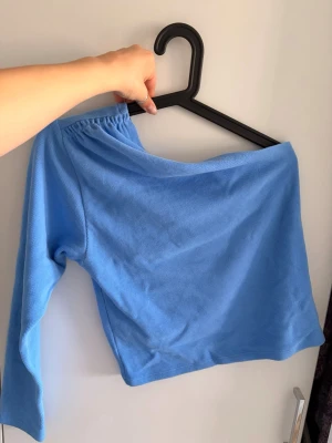 Blå oneshoulder topp från Gina Tricot - Säljer en ljusblå oneshoulder topp från Gina Tricot. Toppen har lång ärm på ena sidan och är axelbandslös på andra sidan. Tillverkad i mjukt ribbat material som sitter snyggt och bekvämt. Perfekt för dig som vill ha en trendig och unik look.