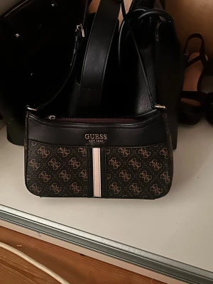 Guess brun monogram handväska - Snygg handväska från Guess i brunt konstläder med klassiskt monogrammönster och en vit samt beige rand framtill. Väskan har guldfärgade detaljer och en smidig axelrem. Perfekt för dig som gillar trendiga accessoarer med logga.