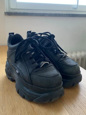 Buffalo London svarta platåsneakers - Buffalo London sneakers i helsvart läder med chunky platåsula och klassisk logga på plösen. Skorna har svarta skosnören, metalliska detaljer och en robust, sportig look som sticker ut. Perfekta för dig som vill ha extra höjd och en edgy stil. Lite slina framåt tårna, enligt bild.