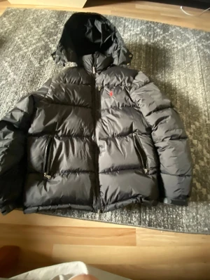 Svart pufferjacka från Polo Ralph Lauren - Snygg svart pufferjacka från Polo Ralph Lauren med huva och dragkedja framtill. Jackan har två stora fickor med dragkedja och den klassiska röda loggan broderad på bröstet. Perfekt för kalla vinterdagar och riktigt skön att bära.