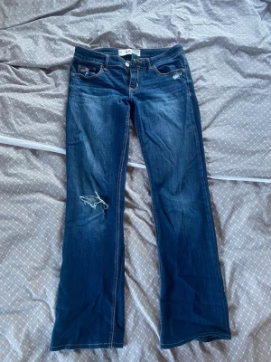 Mörkblå bootcut jeans från Hollister - Snygga mörkblå bootcut jeans från Hollister med slitning på vänster knä och dekorativa sömmar på bakfickorna. Klassisk femficksmodell med låg midja och knappgylf. Perfekta för en avslappnad och trendig look. Uppsydda för att passa min längd (160 cm). 