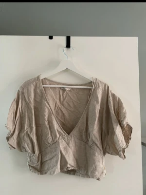Beige croppad linnetopp med v-ringning - Trendig beige croppad topp i linne med djup v-ringning och korta puffärmar. Toppen har en avslappnad passform och är perfekt för varma dagar. Snygg detalj med lätt rynk vid ärmslut och midja. Enkel att matcha med både jeans och kjol.