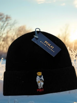 Svart mössa Polo Ralph Lauren - Svart ribbstickad mössa från Polo Ralph Lauren med uppvikt kant och broderad björn i astronautdräkt framtill. Klassisk och stilren design med Polo-logga i rött under björnen. Perfekt accessoar för kalla dagar.