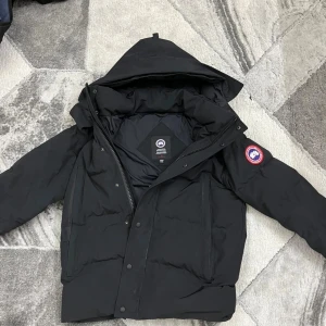 Svart dunjacka från Canada Goose - Säljer en svart dunjacka från Canada Goose med avtagbar huva . Jackan har en klassisk design med dragkedja och knappar framtill samt en ikonisk logga på ärmen. Perfekt för kalla vinterdagar. 