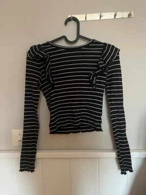 Svartvit randig topp från Kappahl - Snygg svart långärmad topp från Kappahl med vita horisontella ränder. Toppen har volangdetaljer på axlarna och en liten rosett framtill. Tillverkad i mjuk, ribbad bomull som sitter tight och bekvämt.