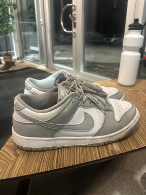 Nike Dunk Low grå och vita sneakers - Nike Dunk Low sneakers i stilren grå och vit färgkombination. Skorna har klassisk låg siluett, vit tåbox och sidor med grå overlays och Swoosh. Ovandelen är i läder och de har platta grå skosnören. Perfekta för en clean och sportig look.