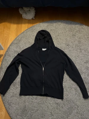 Svart zip hoodie med huva - En enkel svart hoodie med dragkedja framtill och huva. Hoodien har långa ärmar och ribbade muddar vid ärmslut och nederkant. Perfekt för en avslappnad stil och funkar till det mesta. Märke och material framgår ej.