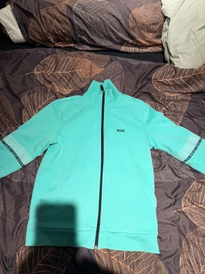 Turkos ziptröja från BOSS & gröna byxor - Säljer en turkos ziptröja från BOSS med svart dragkedja och logga på bröstet samt detaljer på ärmarna. Till det ett par gröna träningsbyxor från Lacoste med resår i midja och benslut, samt liten logga på höften. Perfekt för sport eller chill.