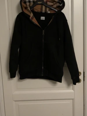 Svart hoodie med Burberry-mönstrad huva - Säljer en svart zip-hoodie från Burberry med klassiskt rutigt foder i huvan. Hoodien har dragkedja framtill, två fickor och ribbade muddar vid ärmar och nederkant. Perfekt för dig som gillar stilrena plagg med en twist.