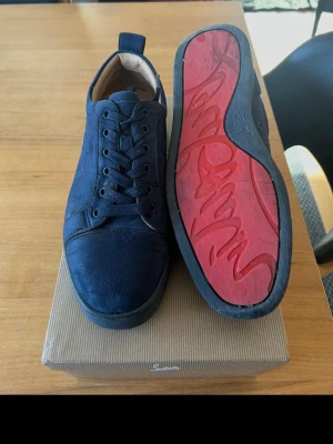Mörkblå sneakers Christian Louboutin - Snygga mörkblå sneakers från Christian Louboutin i mocka med klassisk röd sula och svarta snören. Låg modell med diskret logga på sulan och stilren design. Perfekta för dig som vill ha något exklusivt och trendigt.