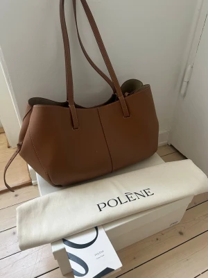 Polene Cyme väska  - Säljer en snygg brun handväska från Polène i mjukt, strukturerat läder. Väskan har långa axelband, minimalistisk design och vita kontrastsömmar. Perfekt för dig som gillar stilrena accessoarer och vill ha plats för allt du behöver.