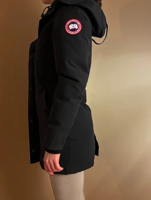 ÄKTA Svart dunjacka från Canada Goose - Snygg svart dunjacka från Canada Goose med huva och klassisk logga på ärmen. Jackan har dragkedja och knappar framtill samt två fickor. Perfekt för kalla dagar och har en stilren, lång modell med lång ärm. Modell: Victoria Parka