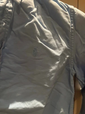 Ljusblå skjorta från Ralph Lauren - Klassisk ljusblå skjorta från Ralph Lauren med broderad logga på bröstet. Skjortan har knappar framtill och krage, samt långa ärmar. Tillverkad i mjuk bomull som känns skön mot huden. Perfekt för en clean och stilren look.