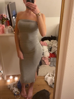 Grå axelbandslös långklänning - Säljer en grå, axelbandslös långklänning i figurnära modell. Klänningen har en slits på sidan och är tillverkad i ett mjukt, stretchigt material som sitter snyggt på kroppen. Perfekt för dig som gillar enkel men stilren design.