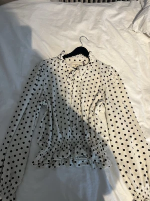 Prickig blus med knyt från Holly & Whyte - Säljer en vit blus med svarta prickar från Holly & Whyte. Blusen har lång ärm, volangdetaljer vid kragen och ett snyggt knyt framtill. Smockad midja och ärmslut ger en trendig siluett. Perfekt för dig som gillar lekfulla mönster och detaljer. 