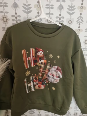 Grön jultröja med tryck - Mysig grön sweatshirt med juligt tryck av snögubbe, tomte och ren samt texten 'Ho Ho Ho' i färgglada bokstäver. Tröjan har rund hals och långa ärmar, perfekt för att sprida julstämning.