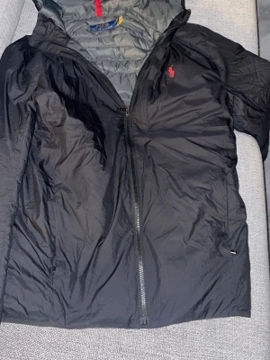 Svart vindjacka från Polo Ralph Lauren - Säljer en svart vindjacka från Polo Ralph Lauren med röd logga på bröstet. Jackan är lätt, har huva och dragkedja framtill. Den är packbar och kan enkelt vikas till en liten påse, perfekt att ta med överallt.