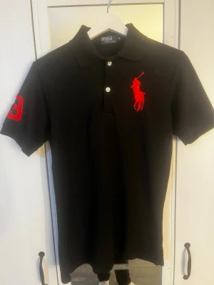 Svart pikétröja från Polo Ralph Lauren - Klassisk svart pikétröja från Polo Ralph Lauren med röd broderad logga på bröstet och röd siffra på ärmen. Tröjan har krage, knappar framtill och korta ärmar. Perfekt till en stilren och sportig look. Har blivit ganska nopprig. Hör av er vid frågor!