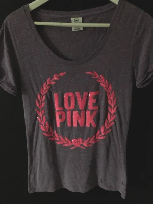 Lila t-shirt från PINK Victoria's Secret - Snygg lila t-shirt från PINK Victoria's Secret med texten 'LOVE PINK' i glittriga rosa bokstäver och en rosa lagerkrans på bröstet. T-shirten har rund halsringning och korta ärmar, perfekt för en avslappnad och trendig look.