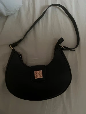 Svart axelväska  - Snygg svart axelväska. guldfärgade metalldetaljer och ett elegant logospänne framtill. Tillverkad i slätt konstläder som ger en stilren look. Knappt använd i nyskick