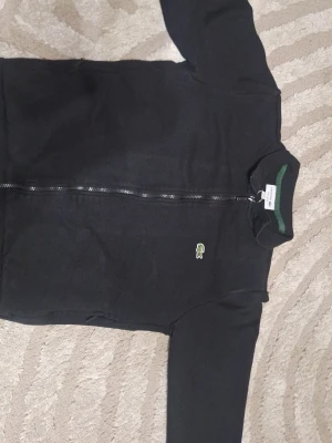 Svart zip tröja från Lacoste - Säljer en svart hoodie från Lacoste med dragkedja framtill och den klassiska gröna krokodilloggan på bröstet. Regular fit och ribbade muddar. Insidan av kragen har en grön detalj. Tillverkad i mjukt bomullsmaterial för skön känsla.