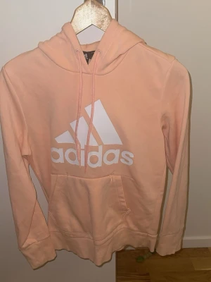 Persikofärgad hoodie från Adidas - Snygg persikofärgad hoodie från Adidas med stor vit logga framtill. Klassisk känguruficka, dragsko i huvan och mjukt material som känns skönt mot huden. Perfekt för en avslappnad och sportig stil.