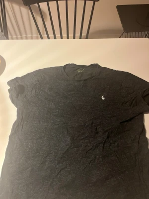 Mörkgrå t-shirt från Ralph Lauren - Snygg mörkgrå t-shirt från Ralph Lauren med klassisk rund hals och den ikoniska vita polospelaren broderad på bröstet. Tillverkad i mjukt bomullsmaterial som känns skönt mot huden. Perfekt basplagg för en clean och stilren look.