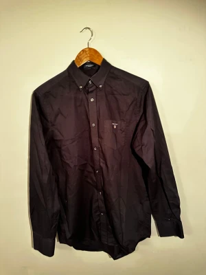 Svart skjorta från Gant med knappar - Klassisk svart skjorta från Gant med button-down krage och bröstficka med diskret logga. Skjortan har långa ärmar och stängs med knappar framtill. Perfekt till en clean och stilren look.