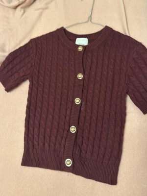 Vinröd kabelstickad kofta H&M - Kortärmad vinröd kabelstickad kofta från H&M med guldfärgade knappar och rund hals. Ribbstickade muddar vid ärmslut och nederkant ger en snygg finish. Perfekt att styla över en topp eller klänning för en trendig look.