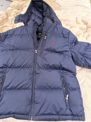 Blå dunjacka från Polo Ralph Lauren - Snygg blå dunjacka från Polo Ralph Lauren med huva och dragkedjor på fickorna. Jackan har en röd logga på bröstet och en extra bröstficka med dragkedja. Perfekt för kalla vinterdagar och riktigt stilren design.i storlek xxl från kidsbrandstore så den är som en S/M i vanliga storlek pris går att diskutera 
