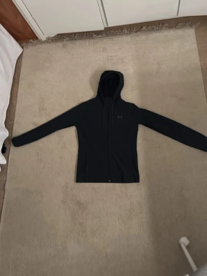 Svart vindjacka från Under Armour - Svart vindjacka med dragkedja från Under Armour. Vindjackan har långa ärmar, justerbar huva med snören och diskret logga på bröstet. Tillverkad i ett mjukt och stretchigt material som passar perfekt för träning eller chill. Storlek S men kan också passa XS