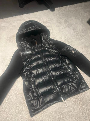 Svart dunjacka med huva från Moncler - Snygg svart dunjacka från Moncler med glansig quiltad kropp, stickade svarta ärmar och huva. Jackan har dragkedja framtill och en ficka med Moncler-logga på ena ärmen. Perfekt för kalla vinterdagar och riktigt stilren look.