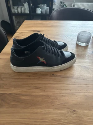 Svarta sneakers från Axel Arigato - Snygga svarta sneakers från Axel Arigato med vit sula och broderad fågel i färg på sidan. Skorna är använda men fortfarande i Okej skick, defekt på båda skorna är att lappen släppt lite runt om kring fågeln vilket går att åtgärda, annars lite mer puts så är dem näst intill som nya! Ny pris ca 2500k, mitt pris 400kr