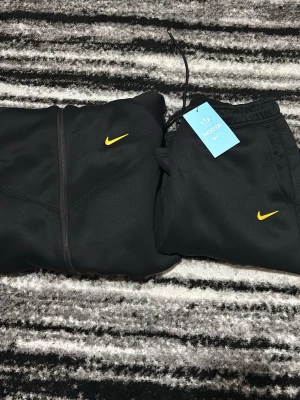 Svart tracksuit från Nike NOCTA - Svart tracksuit från Nike NOCTA med gul swoosh på bröstet och dragkedja framtill. Hoodien har huva med dragsko och två fickor med dragkedja. Perfekt för en sportig och stilren look.