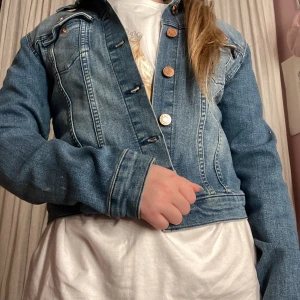 Klassisk blå jeansjacka - Snygg blå jeansjacka med kopparfärgade knappar och två bröstfickor. Figurnära passform och långa ärmar. Perfekt att slänga över en t-shirt för en avslappnad look. Tidlös modell som passar till det mesta.