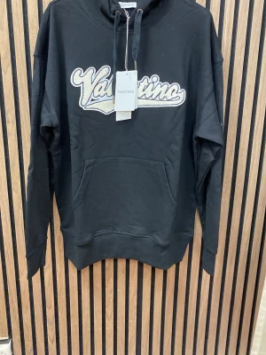 Svart hoodie  - Snygg svart hoodie  med stor broderad logga i vitt och beige på bröstet. Klassisk känguruficka framtill, ribbade muddar och justerbar huva med snören. Perfekt för en avslappnad och trendig look.