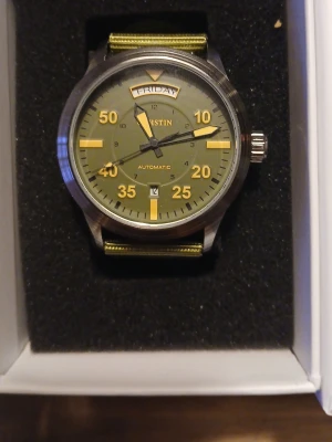 Grön automatisk klocka från Austin - Snygg automatisk klocka från Austin med grön urtavla och matchande grönt nylonband. Urtavlan har tydliga gula siffror och markeringar, dag- och datumvisning samt robust boett i metall. Perfekt för dig som gillar en sportig och stilren look.