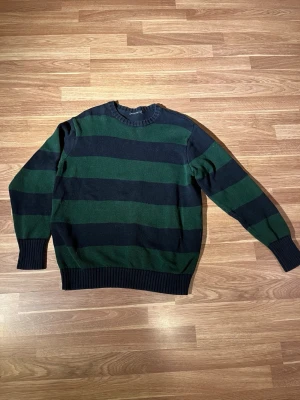 Grön och svart randig stickad tröja - Stickad tröja med breda gröna och svarta ränder. Klassisk rund halsringning och ribbade muddar vid ärmslut och nederkant.