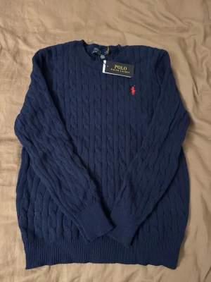 Mörkblå kabelstickad tröja Polo Ralph Lauren - Kabelstickad mörkblå tröja från Polo Ralph Lauren med rund halsringning och röd broderad logga på bröstet. Tröjan har långa ärmar och ribbade muddar vid ärmslut och nederkant. Perfekt för dig som gillar klassisk och stilren look.
