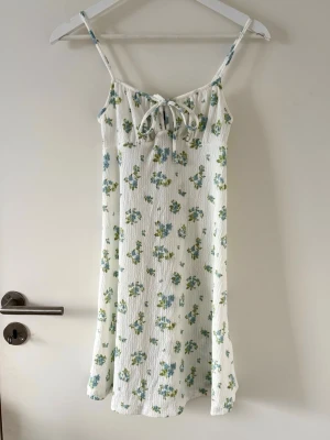 Vit blommig kortklänning  - Supersöt vit kortklänning från Gina Tricot med blå och gröna blommor, smala axelband och knyt fram. 